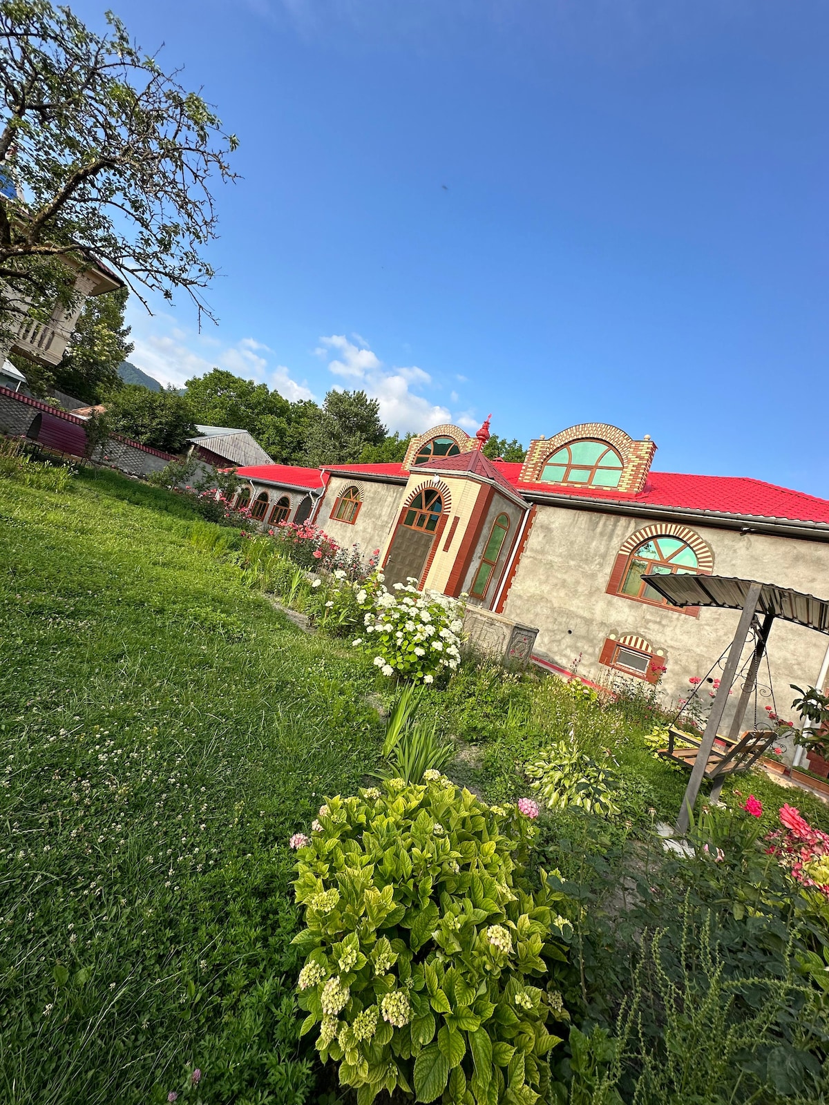 A quaint exterior of a house is visible, showcasing a vibrant red roof and decorative windows. Lush greenery surrounds the structure, with a variety of flowering plants and shrubs in the foreground. A clear blue sky adds to the serene atmosphere.