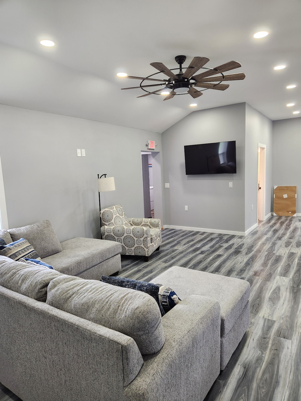 A spacious living area features a light gray sectional sofa and a patterned armchair. Recessed lighting and a ceiling fan provide illumination and airflow. A wall-mounted TV is positioned across from the seating, enhancing the functionality of the space.