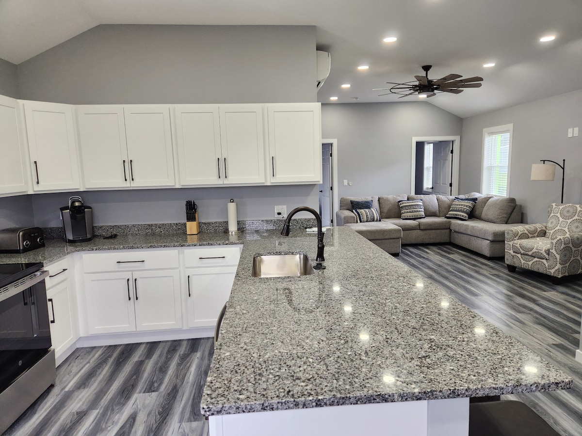 A modern kitchen featuring ample granite countertop space includes a sink and a stove. White cabinetry and stainless-steel appliances provide a clean look. The open layout connects to a cozy living area, which contains a spacious sofa and a floor lamp, inviting relaxation.
