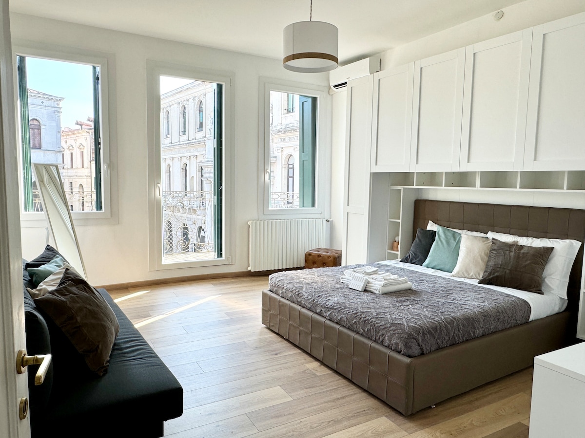 The spacious bedroom features large windows allowing ample natural light, offering views of the historic architecture outside. A comfortable bed with layered bedding is complemented by a cozy seating area and modern furnishings, creating an inviting and restful environment.