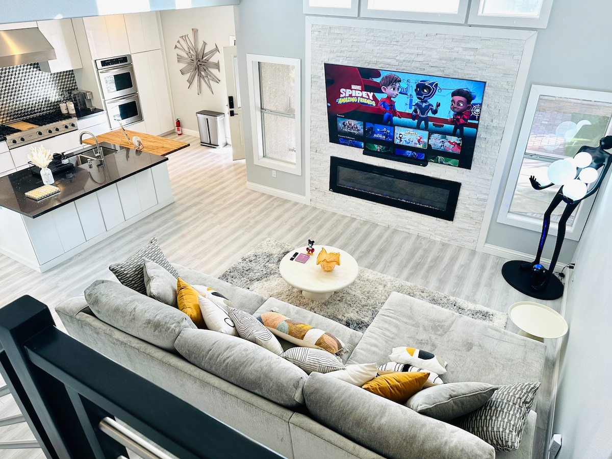 The living room and kitchen area are visible from above, featuring a spacious sofa adorned with decorative pillows. An 80-inch smart TV is mounted above a sleek fireplace. A modern kitchen is equipped with stainless steel appliances and a large island. Natural light fills the open space.