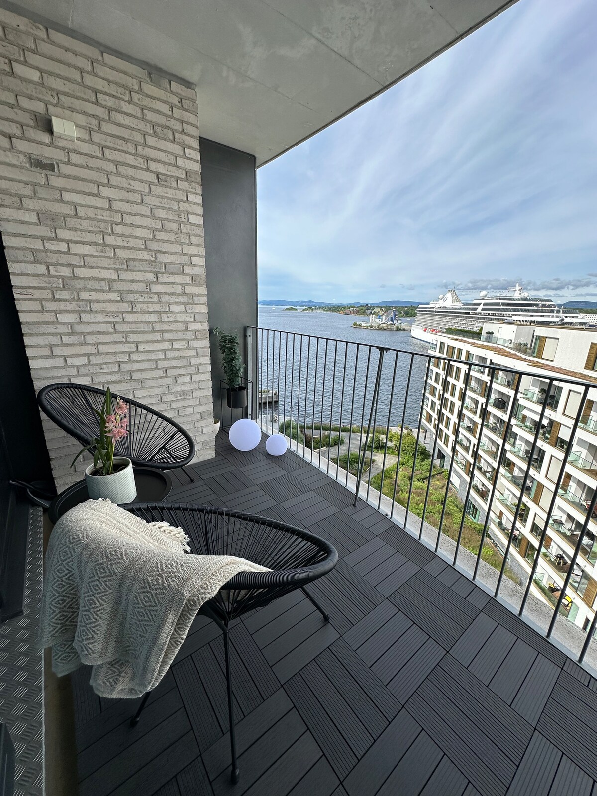 A balcony is equipped with two black wicker chairs and a small table. A decorative plant is positioned beside one chair. A cozy blanket is draped over the railing. The view captures the serene waters of the Oslofjord and modern buildings in the foreground.