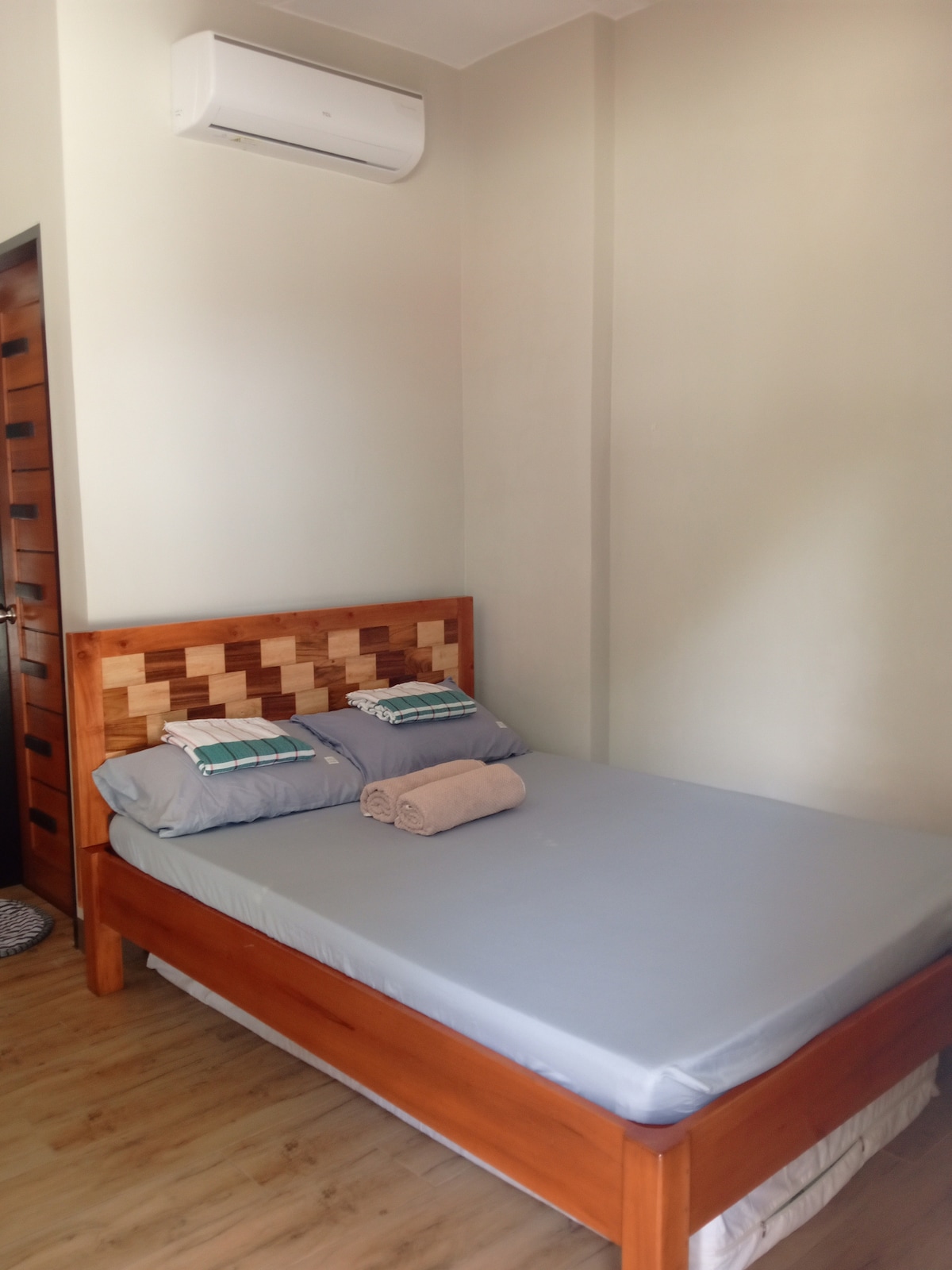 A clean sleeping area features a queen-sized bed with a wooden headboard, accentuated by soft blue and white pillows. A rolled towel is placed neatly on the bed. Natural light illuminates the space, and an air-conditioning unit is mounted above.