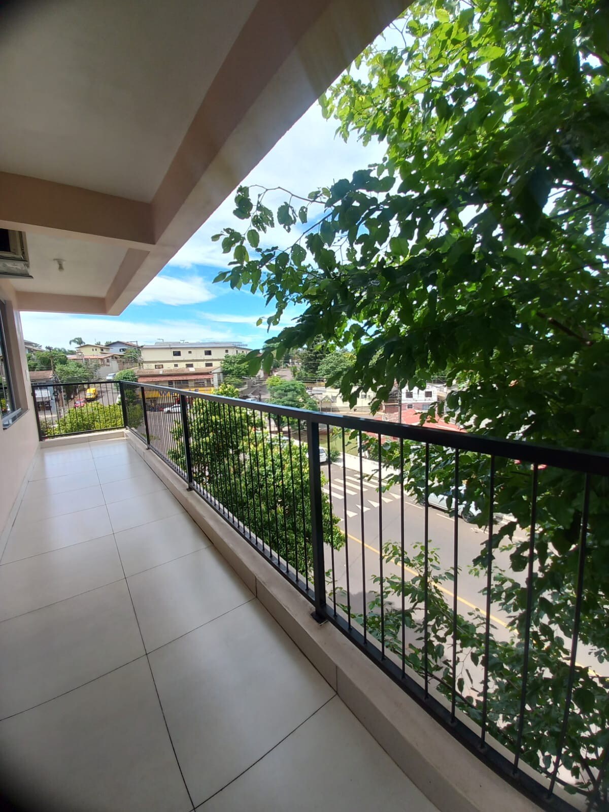 An inviting balcony space is bordered by a black metal railing, offering views of the lush greenery and nearby streets. Bright natural light fills the area, highlighting the tiled floor and the serene atmosphere created by the surrounding trees.