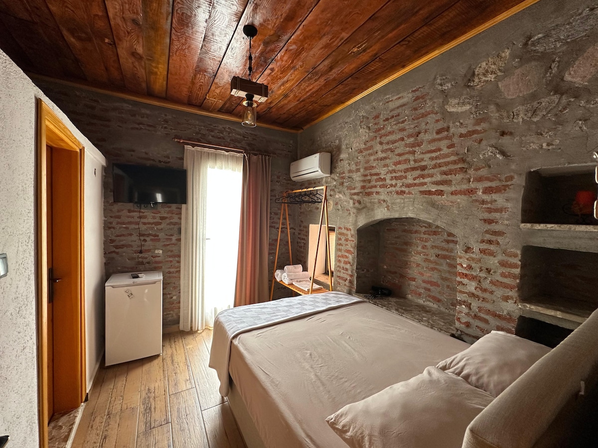 A cozy room features exposed brick walls and wooden ceiling beams. A spacious bed is centered, with light filtered through curtains. A small refrigerator is visible, along with open shelving and a drying rack, creating an inviting and functional space for relaxation.