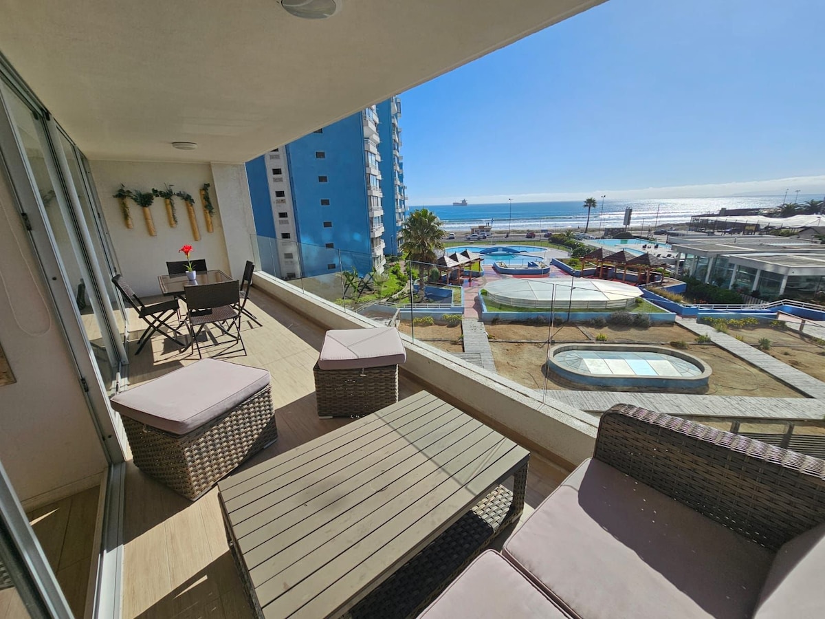 A spacious balcony features outdoor seating, including a table with chairs and lounge furniture. The view overlooks a vibrant area with pools and recreational spaces, while the ocean is visible in the background, creating a serene outdoor experience.