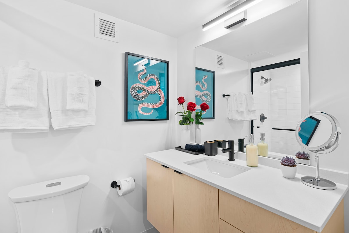 A modern bathroom features a clean white aesthetic with a spacious vanity. Fresh red roses are displayed on the counter beside various toiletries, including candles and a small plant. Bright towels hang neatly on a rack, and a mirror illuminates the space.