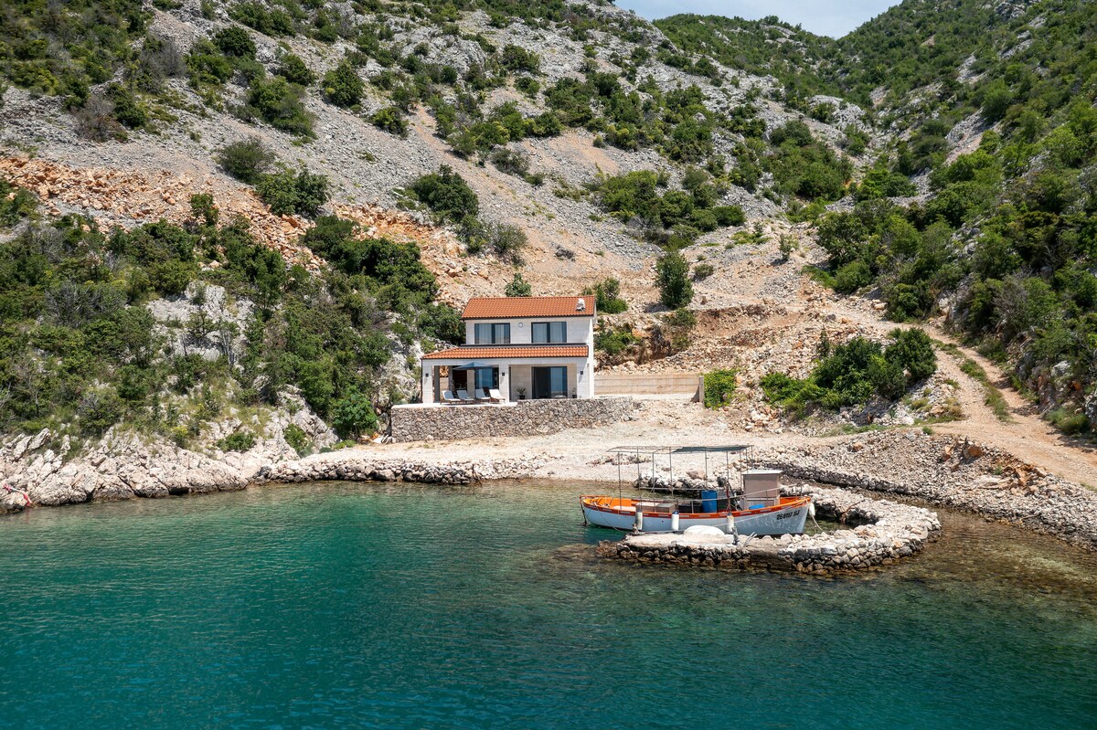 A secluded house is set against a mountainous backdrop, positioned alongside a clear blue sea. A small boat rests at the rocky shoreline, while the home features a modest roof and stone walls, creating a serene environment embraced by nature.