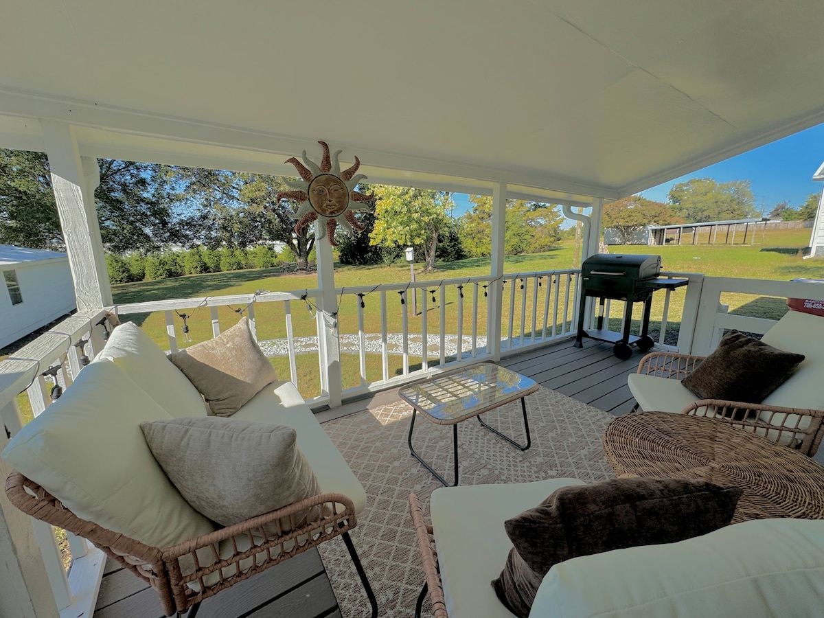 The covered porch features comfortable seating with soft cushions, a small table, and a grill for outdoor cooking. Sunlight filters through the open space, highlighting the serene view of the landscaped yard and greenery beyond.