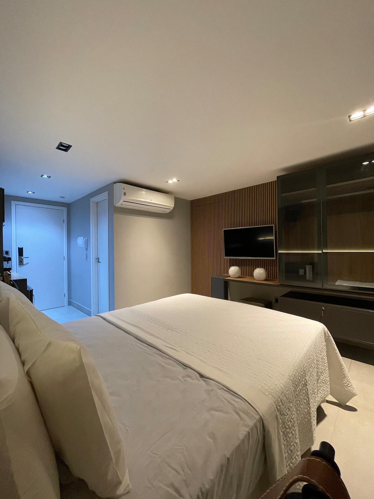 A serene bedroom features a spacious bed covered with a light-colored blanket. A sleek cabinet and a flat-screen television are mounted on the wall, while modern lighting fixtures softly illuminate the room. The air conditioning unit is visible, contributing to a comfortable environment.