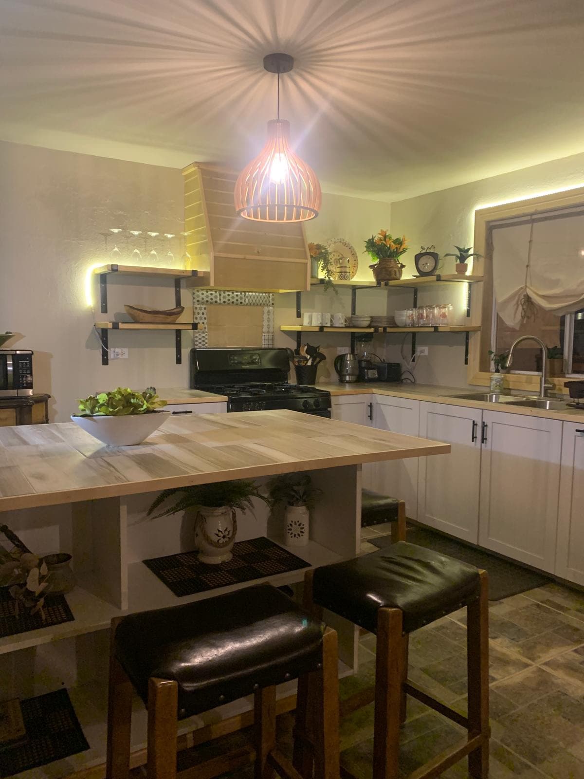 The kitchen features modern cabinetry and a wooden island with seating for four. A warm light fixture hangs above, illuminating the countertop. Shelves are lined with decorative items, and essential kitchen appliances are arranged for convenience.