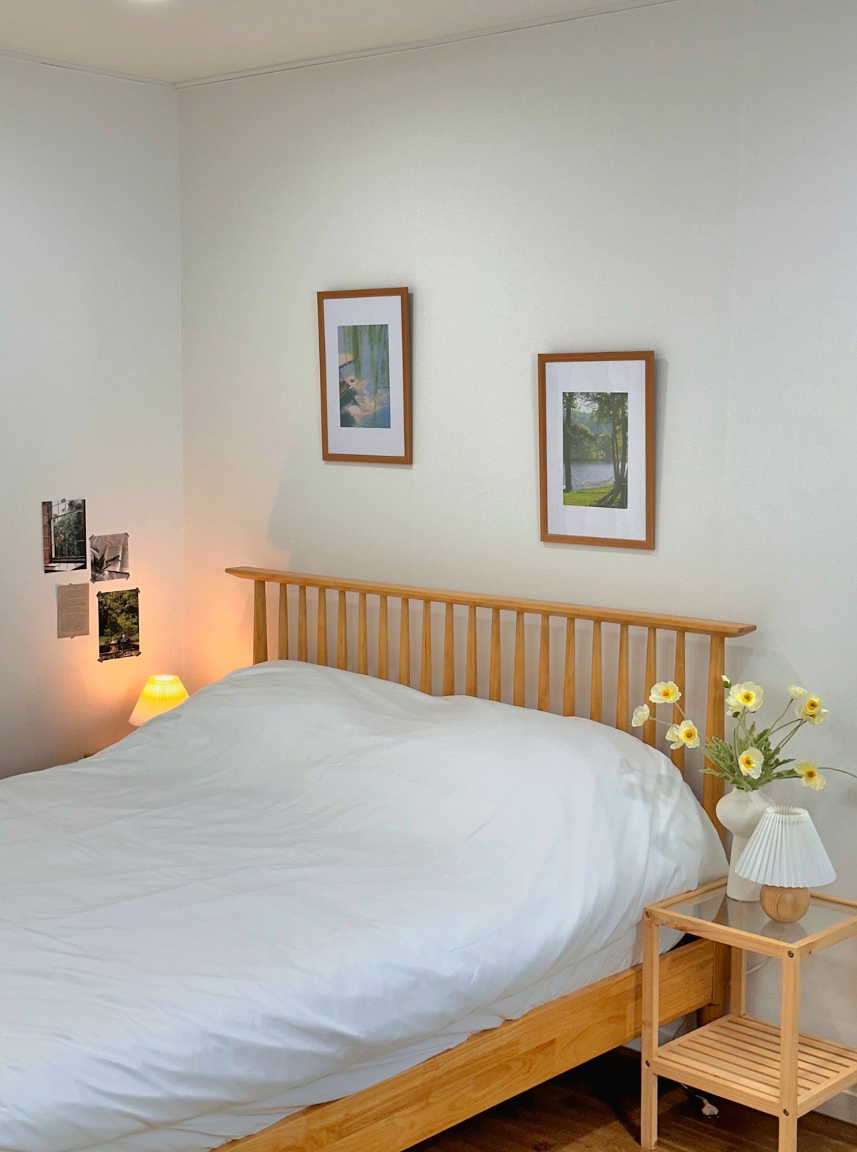 A cozy sleeping space features a spacious bed with white bedding. Wooden frames hold two pictures on the wall, while a small lamp adds soft illumination. A vase with fresh flowers is placed on a bedside table, enhancing the minimalistic decor.