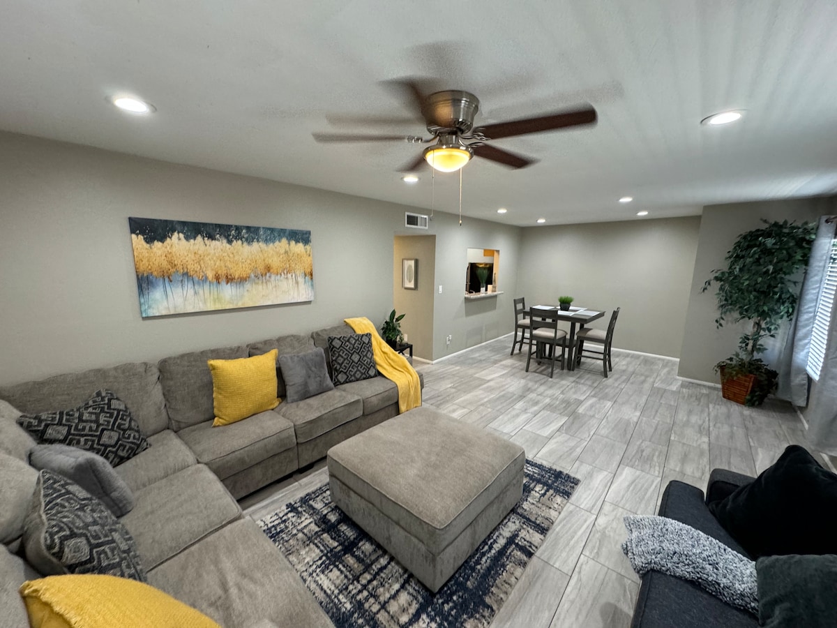 A modern living room features a large sectional sofa, accented with yellow and patterned throw pillows. A cozy area rug is positioned under a central coffee table. Bright overhead lights illuminate the space, which includes a dining table in the background and a wall art piece displaying soothing colors.