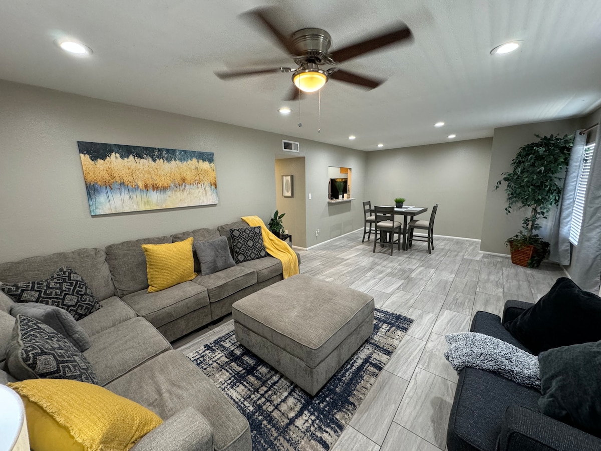 A spacious living area features a large sectional sofa with decorative yellow and patterned pillows. An ottoman sits in the center, paired with a dining table and chairs in the background. Natural light enters through multiple windows, illuminating the neutral-toned walls and tile flooring.