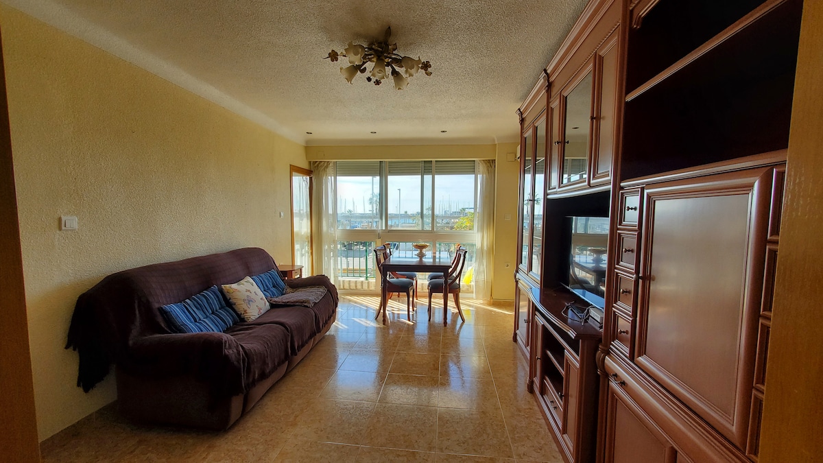 A spacious living area showcases a comfortable sofa alongside a dining table with four chairs. Large windows allow natural light to fill the room, offering views of the surroundings. A built-in entertainment unit adds functional storage, while the soft lighting creates a warm environment.