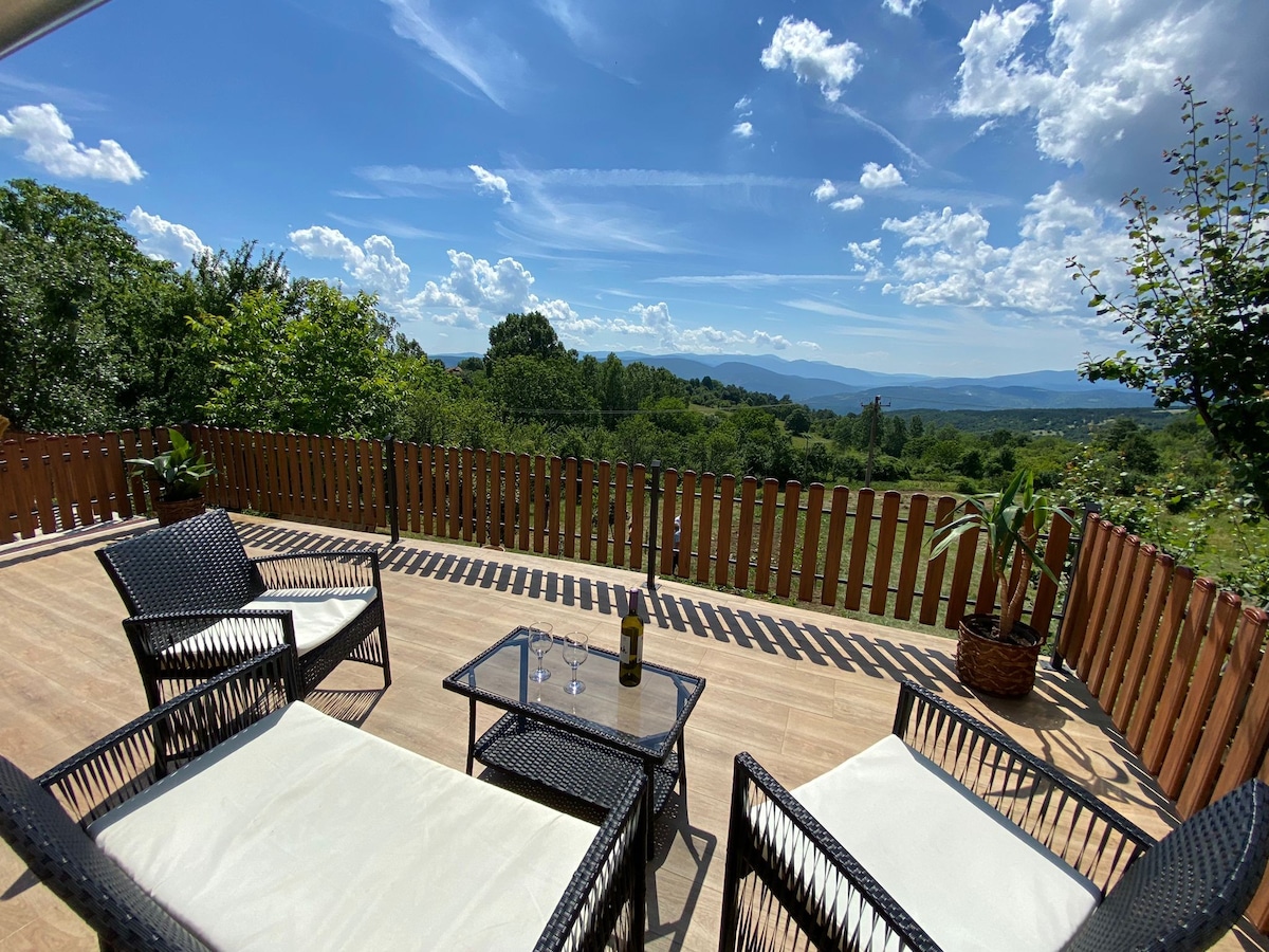 An outdoor terrace is presented with two wicker chairs and a glass table, set against a backdrop of rolling hills and a clear sky. Lush greenery is visible in the surrounding landscape, and a bottle and glasses are arranged on the table.