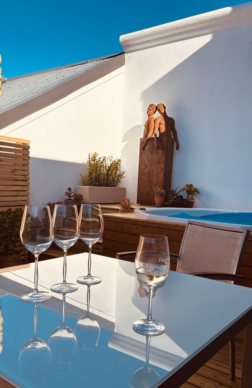 A sunlit outdoor deck features a wooden table with four elegant wine glasses arranged on it. In the background, a private jacuzzi is visible, accompanied by a modern sculpture depicting two figures. Lush greenery enhances the tranquil atmosphere.