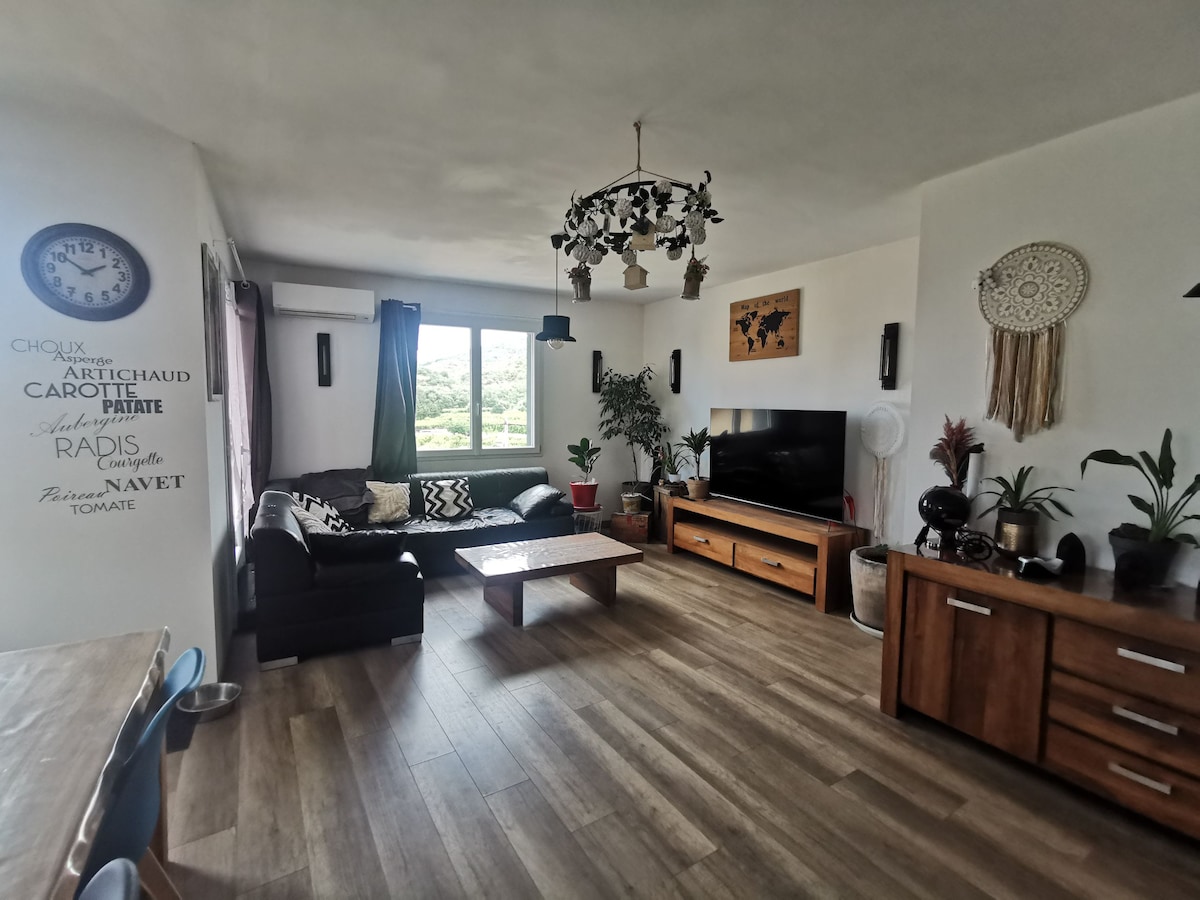 A spacious living room features large windows allowing ample natural light to fill the area. A comfortable dark sofa faces a flat-screen television. Indoor plants are arranged thoughtfully, and a wooden sideboard adds warmth to the space. Decorative elements, including a wall clock and wall hangings, enhance the decor.