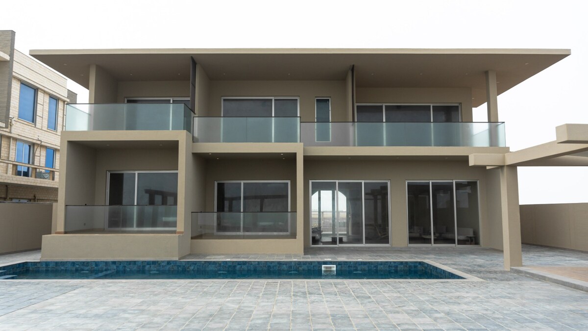 A modern villa features a spacious outdoor terrace and balconies above, providing views of the surroundings. Large glass doors and windows enhance the open design, with a private swimming pool situated in front on a tiled surface.