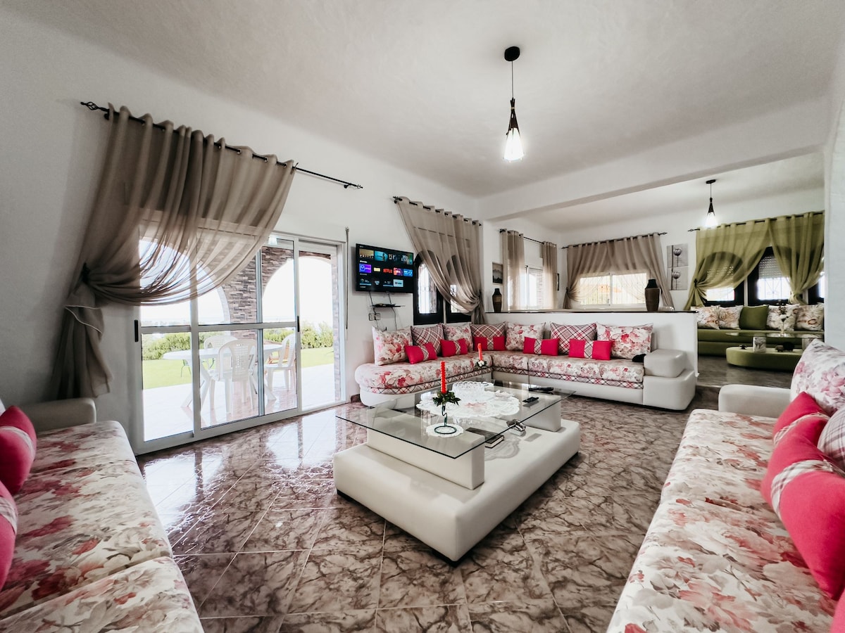 A spacious living area showcases soft floral patterned sofas adorned with bright pink cushions. A glass coffee table sits in the center, surrounded by natural light streaming through large windows with sheer curtains. TV is mounted on the wall, creating a cozy, family-friendly atmosphere.