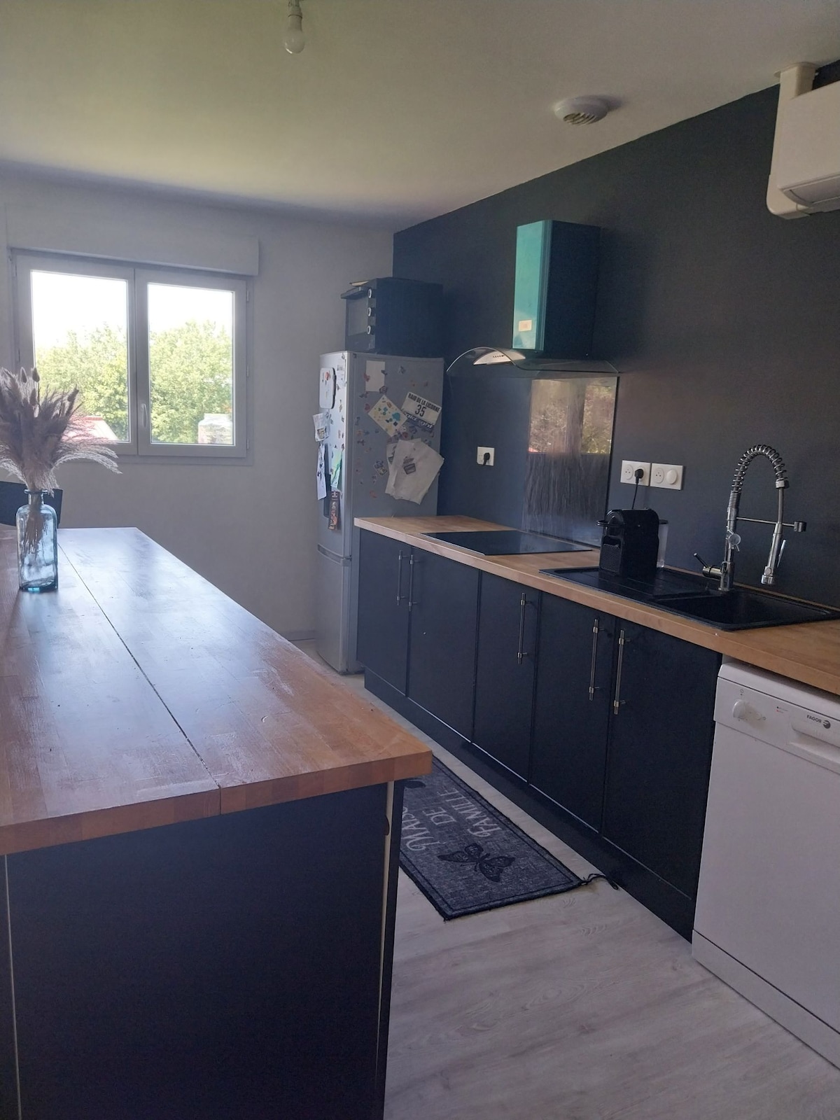A modern kitchen is highlighted with dark cabinetry and wooden countertops. Appliances include a refrigerator and a dishwasher. A sink with a sleek faucet is positioned beside an ample workspace. Natural light enters through a large window, illuminating the space's clean lines.