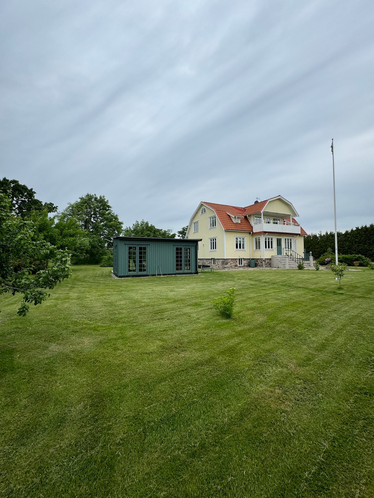 A spacious yard surrounds a two-story yellow house with a red roof, featuring a modern green shed. Neatly trimmed grass highlights the outdoor space, while trees are positioned around the perimeter, adding a natural touch to the setting.