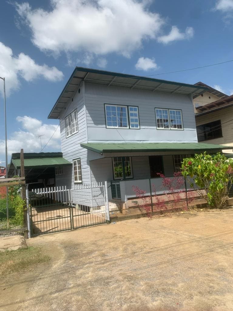 A two-story wooden house with a green metal roof and a welcoming front porch is visible. Large windows allow natural light to flow into the living spaces. A well-maintained garden with flowering plants and a gravel pathway leads to the entrance.