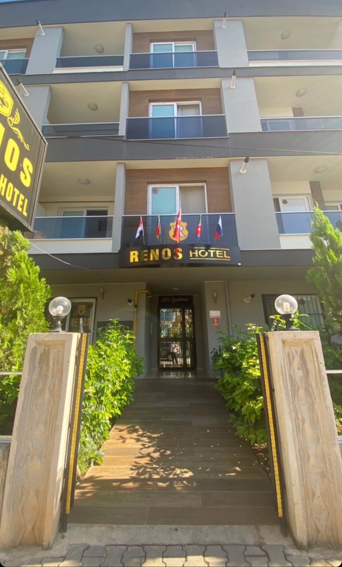 A modern hotel entrance is flanked by neatly trimmed greenery. The entrance features a welcoming sign displaying the hotel name and several flags. Stone steps lead up to the lobby, which is visible through large glass doors.