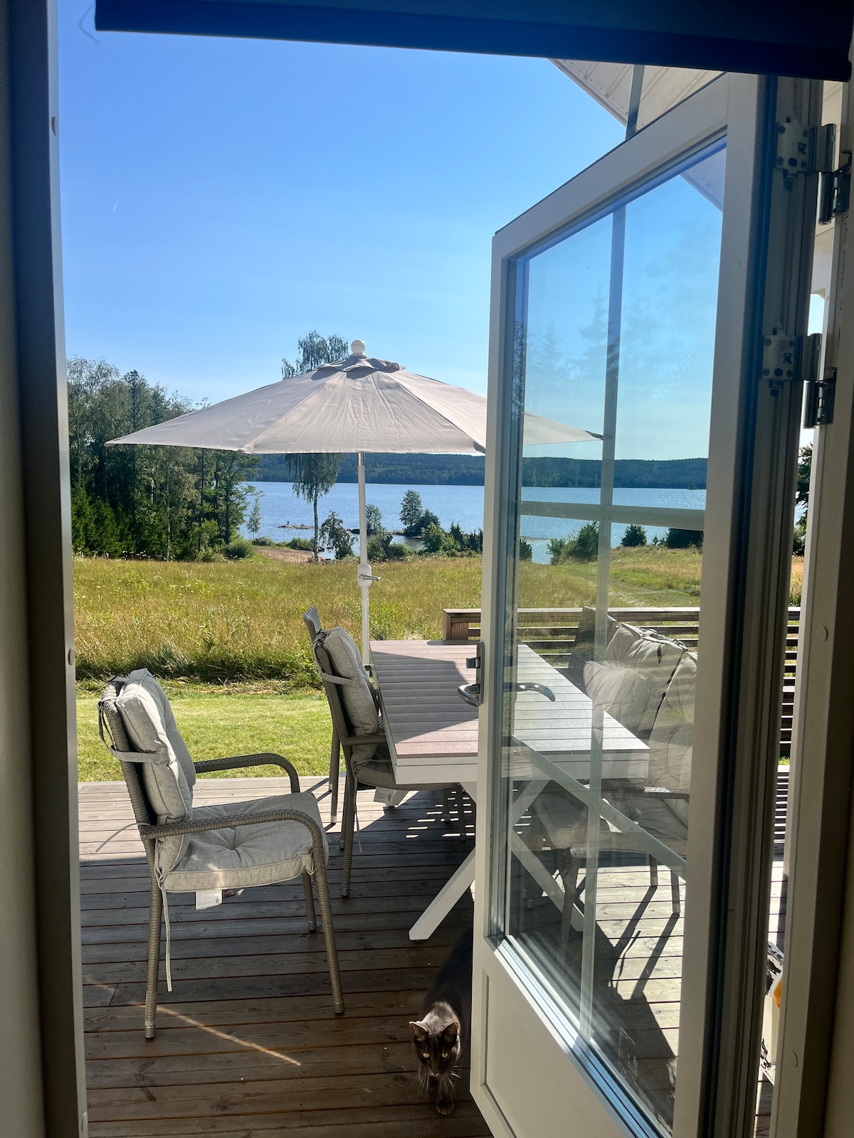 A view from an open door reveals a spacious wooden deck with a dining table and several chairs shaded by a large umbrella. Across the deck, the serene lake and surrounding greenery are visible under a clear blue sky, creating a tranquil outdoor setting.