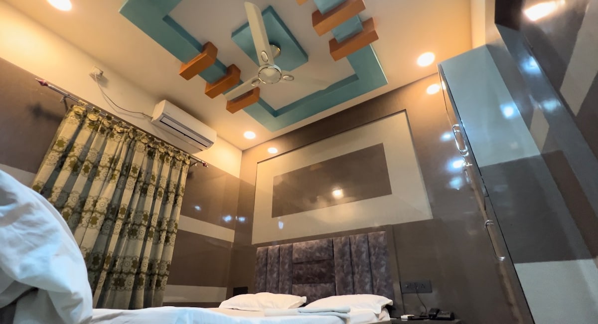 A well-lit room showcases a contemporary design with a uniquely patterned ceiling and elegant wall details. Two spacious beds are neatly arranged, with crisp white linens. Decorative curtains frame a window, while a ceiling fan contributes to a comfortable atmosphere.