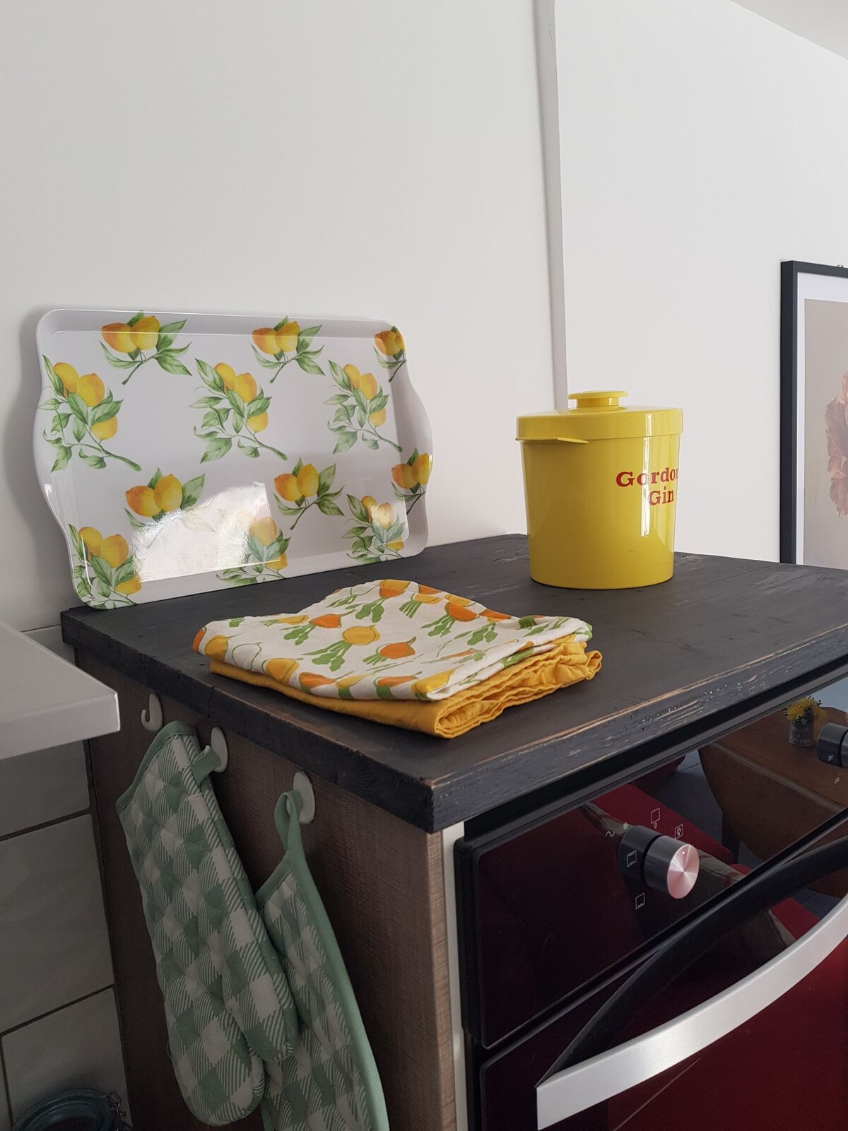 A kitchen countertop is displayed with a decorative tray featuring a lemon pattern, accompanied by a yellow container. A folded towel with an orange design and a green checkered towel hang nearby, adding vibrant colors to the functional space.
