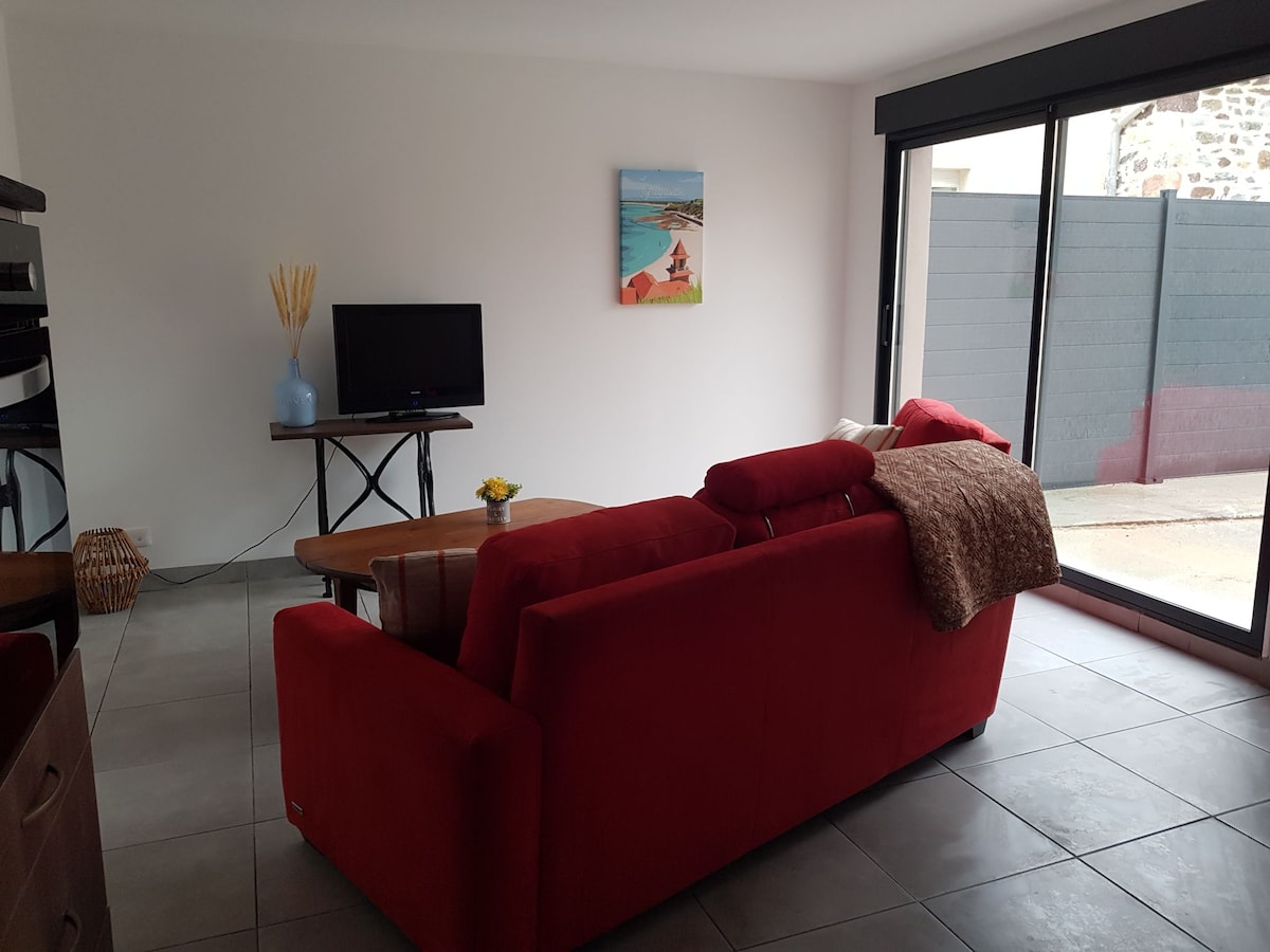 A bright living space is captured, featuring a red sofa positioned near a wooden coffee table. A television is placed on a stand, and a small vase with decorative elements is visible on the table. Glass sliding doors open to a small, enclosed courtyard.