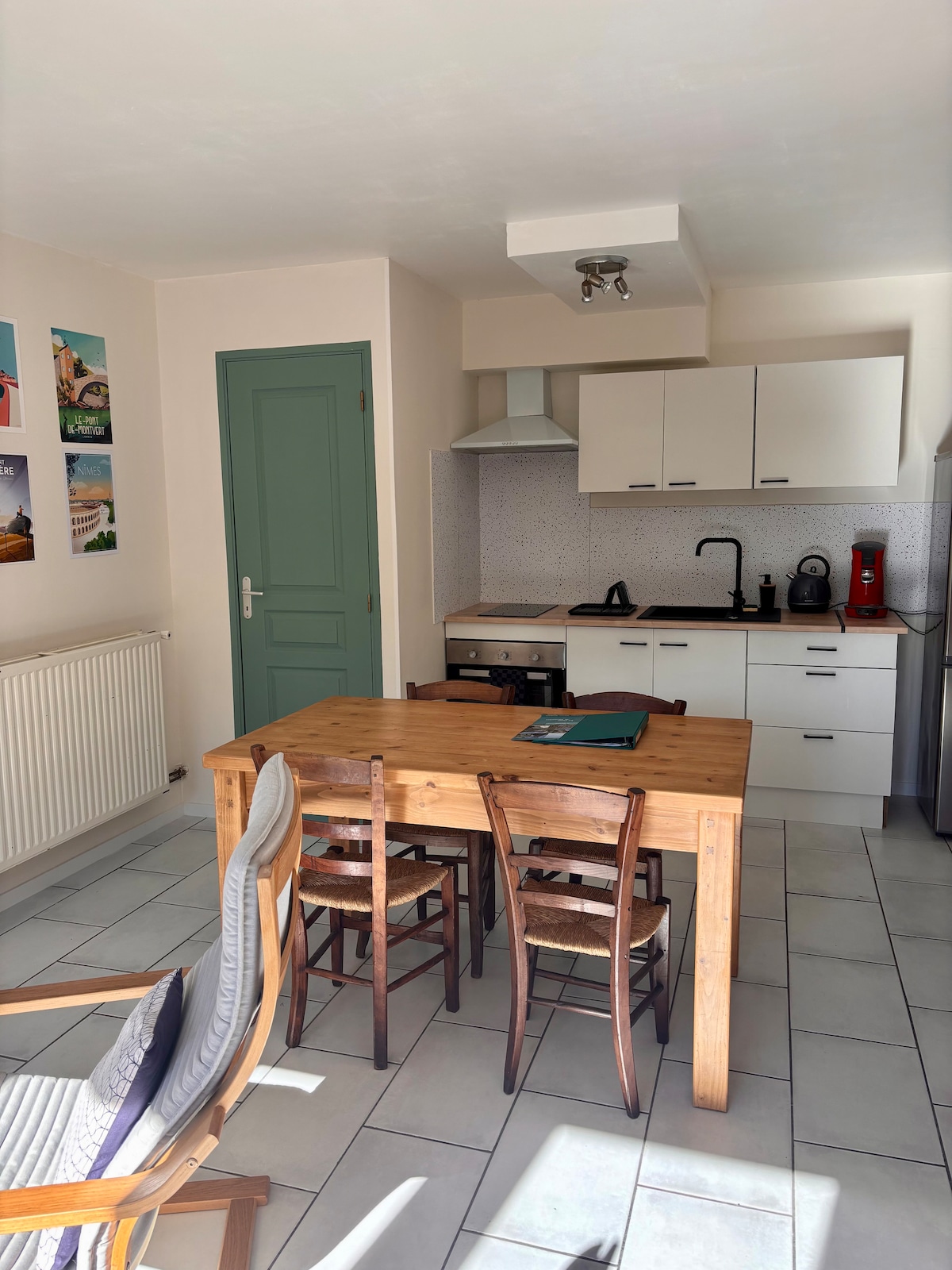 A well-equipped kitchen area is featured, showcasing a wooden dining table surrounded by four chairs. Cabinets line the walls, along with a stove and kitchen appliances. Natural light fills the space, enhanced by a wall decorated with scenic images.