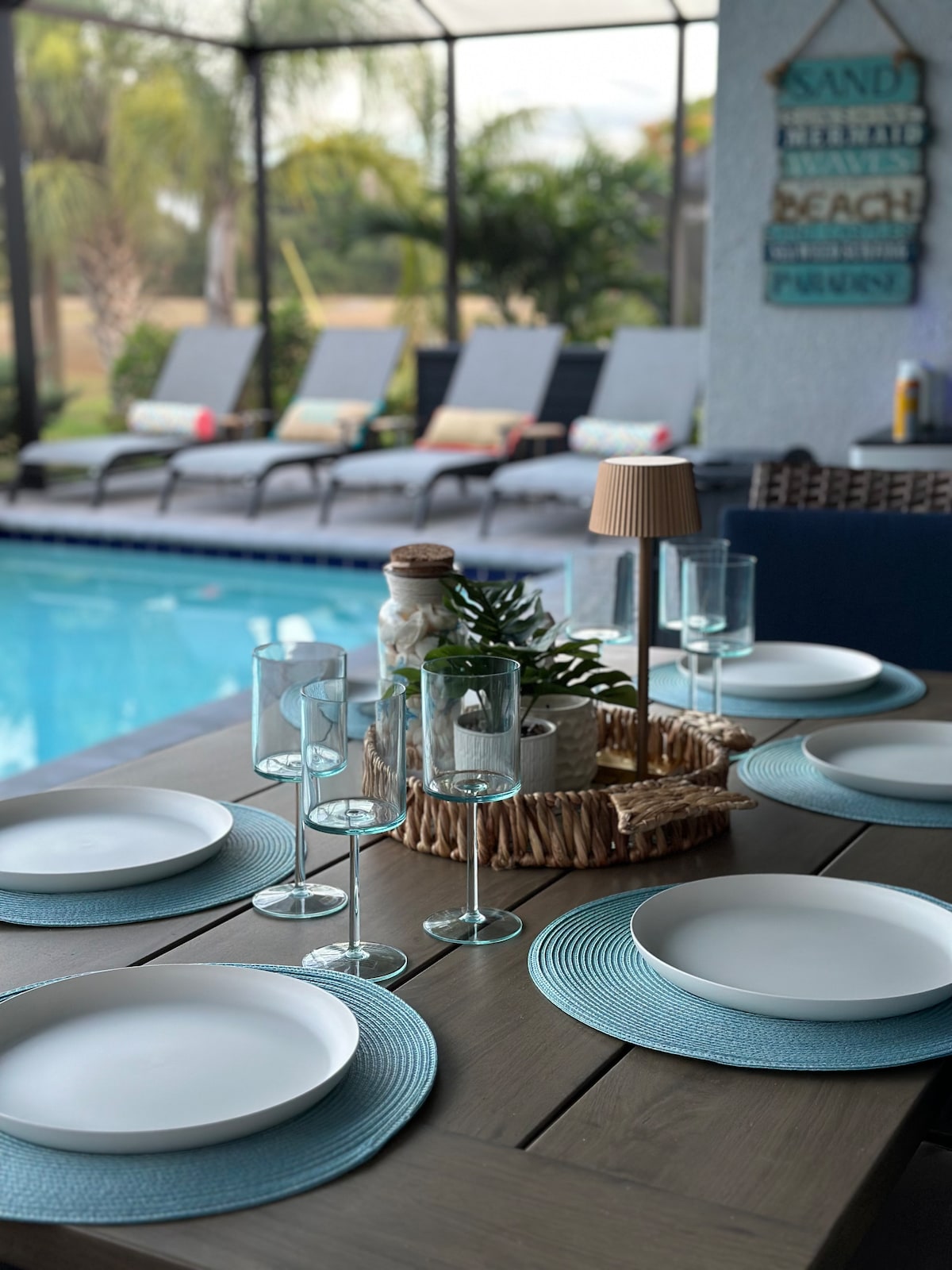 An outdoor dining table is set with light blue placemats and white plates, accompanied by clear glassware. A decorative basket with a plant centerpiece and a small lamp adds a touch of charm. The background features a serene pool and lounge chairs, surrounded by lush greenery.