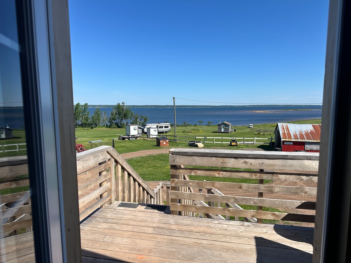 A view from the second-story balcony deck showcases a scenic landscape, including a tranquil body of water and grassy areas. RV sites and rustic buildings are visible in the foreground, framed by clear blue skies.