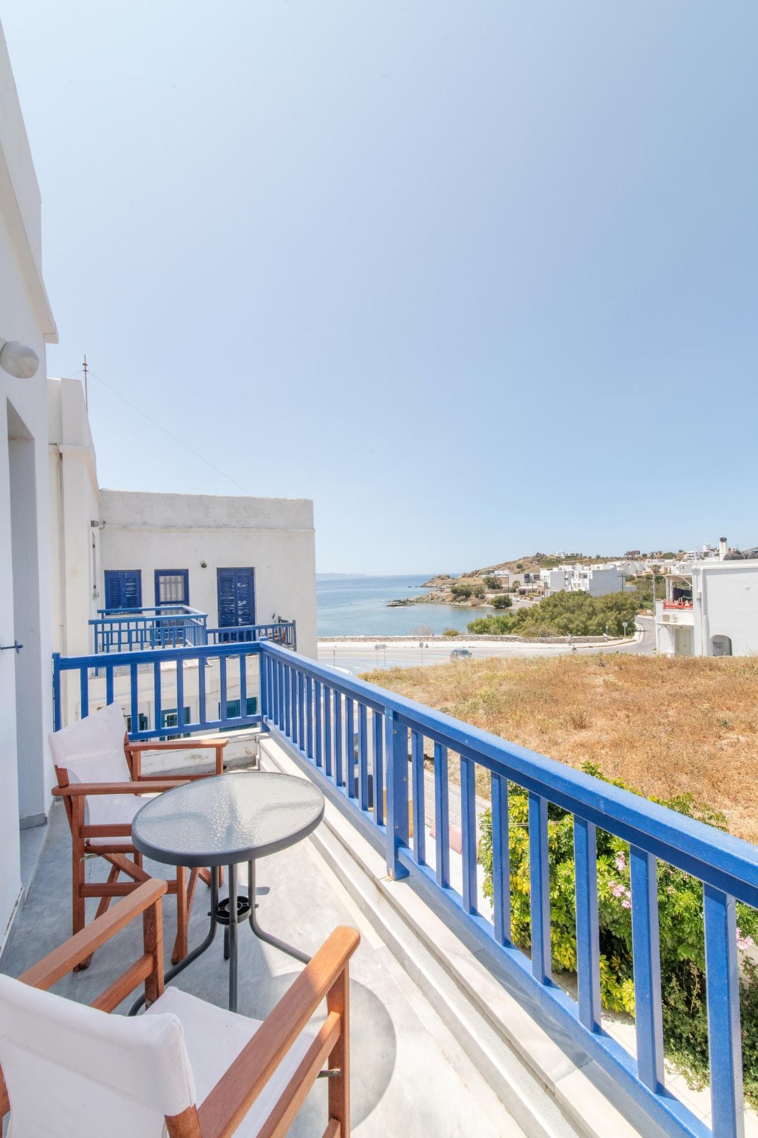 A balcony features blue railings, offering a view of the sea and surrounding landscape. Two wooden chairs and a small round table are visible, creating a cozy outdoor seating area perfect for relaxation.