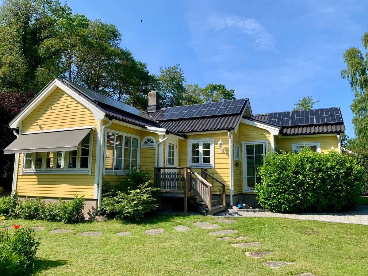 A charming yellow house is framed by lush greenery and a clear blue sky. Solar panels are positioned on the roof. Stone walkways lead up to the entrance, which features a wooden deck and large windows that invite natural light.