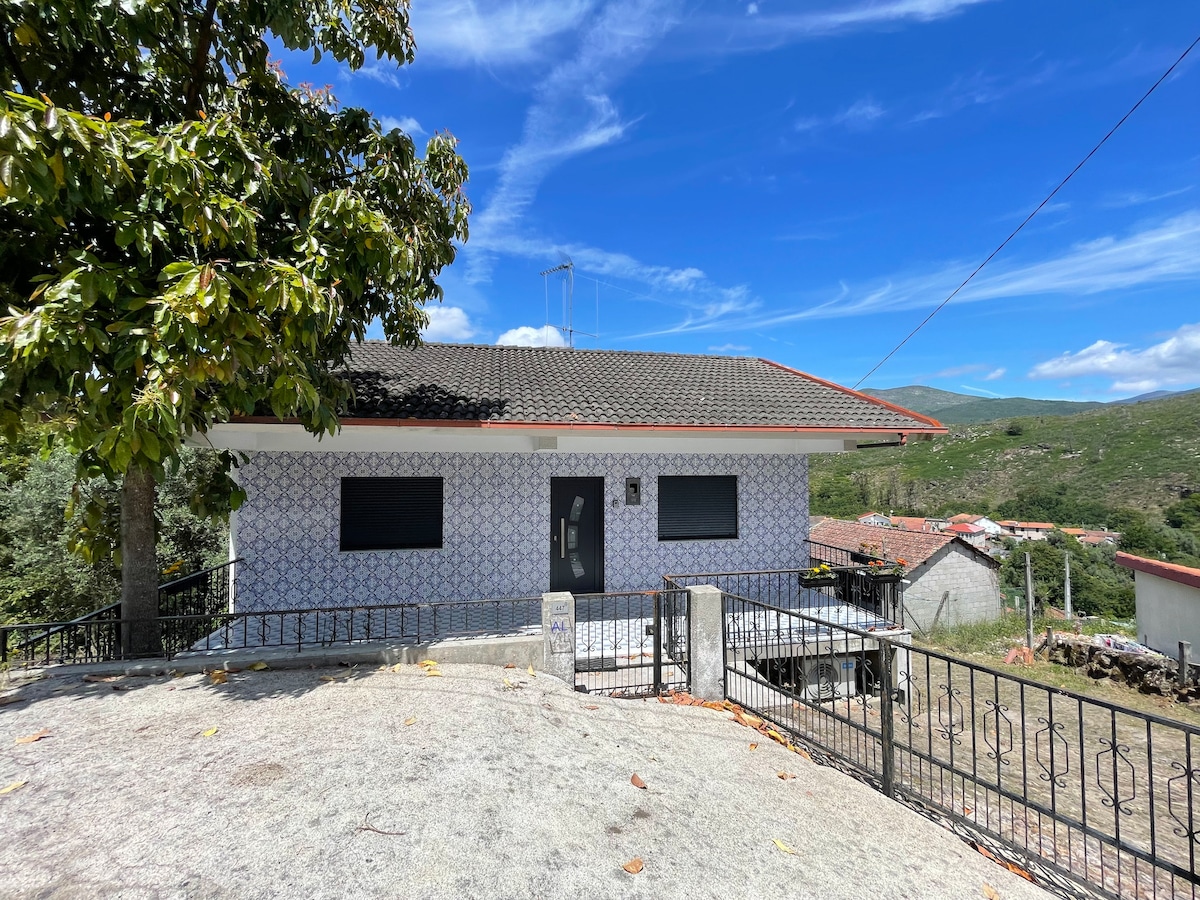 A traditional house is set on a rocky elevation, featuring a patterned exterior and large windows. A stone terrace with a low railing extends in front of the house, offering views of the surrounding landscape and distant hills under a clear blue sky.