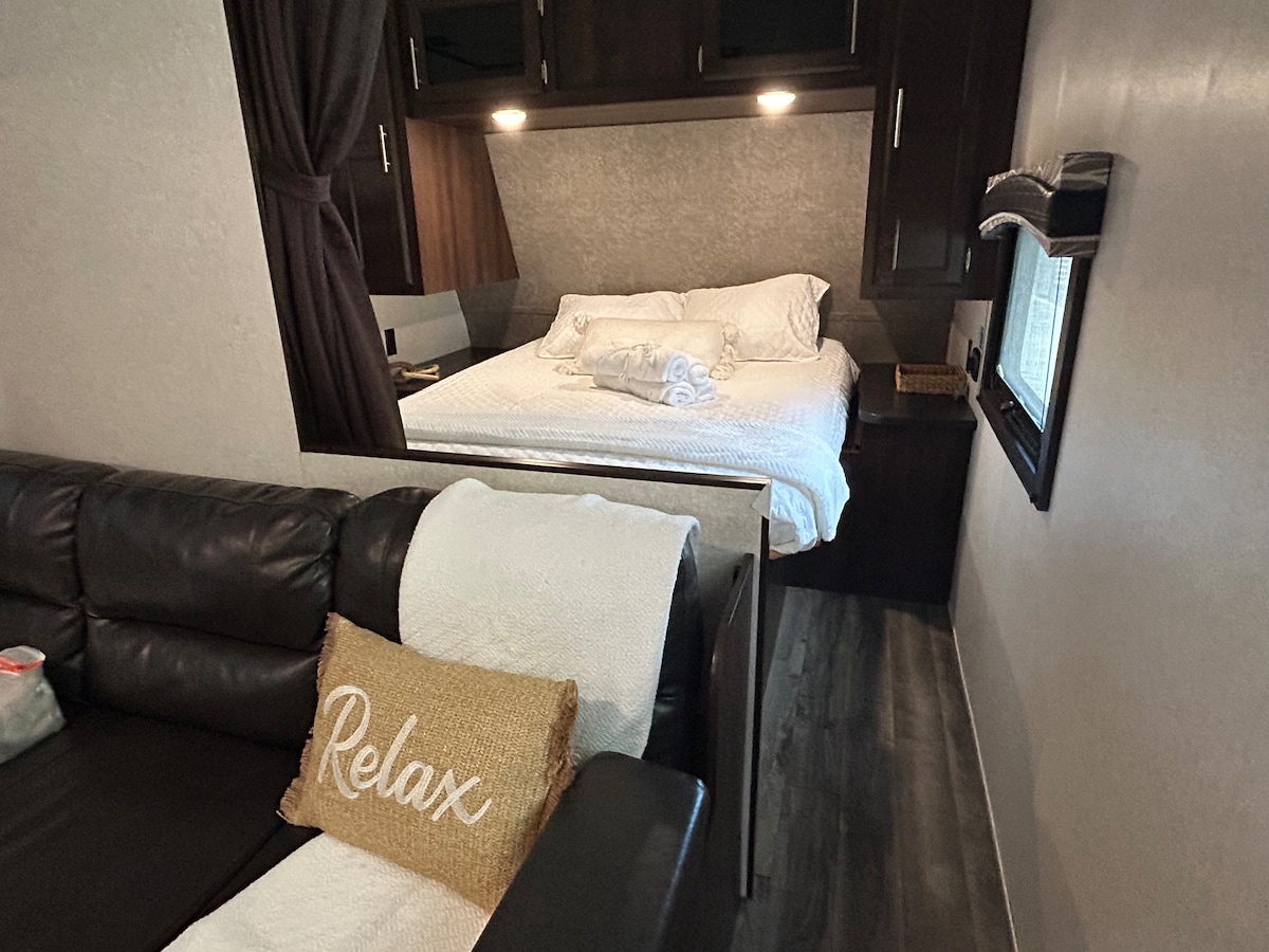 A compact living space displays a spacious bed with neatly arranged white linens and towels. A comfortable dark leather couch, adorned with a decorative 'Relax' pillow, invites relaxation. Natural light filters through a window, highlighting the modern decor and calming color palette.