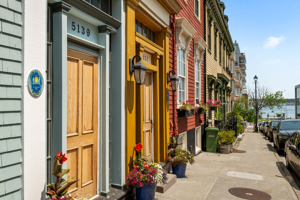 A vibrant row of historic townhouse entrances, featuring a mix of colors from soft blue to warm yellow. Flower-filled planters adorn each doorway, adding natural beauty to the scene. A view of the waterfront is visible in the background under a clear sky.