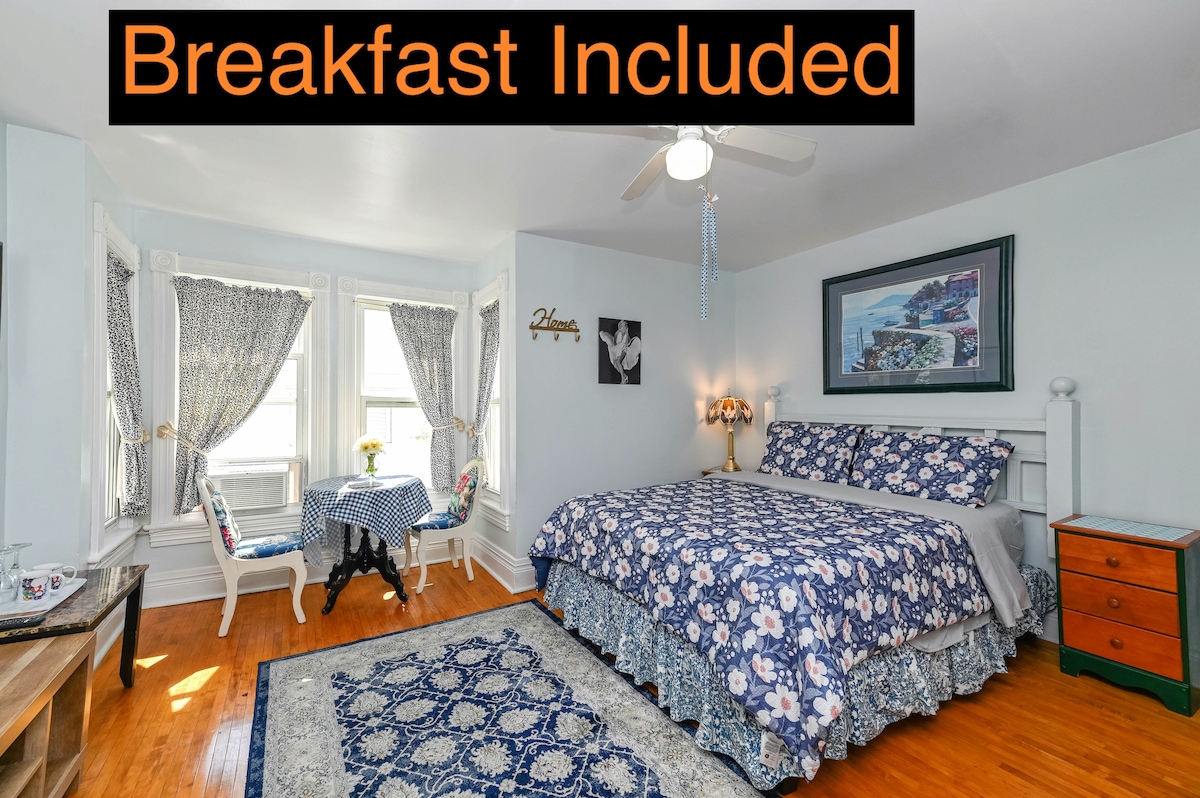 A guest room features a spacious bed covered in floral bedding, complemented by a decorative rug. A small dining table with checkered tablecloth and two chairs is situated near the windows, providing natural light. A bright, inviting atmosphere is created by the light blue walls.