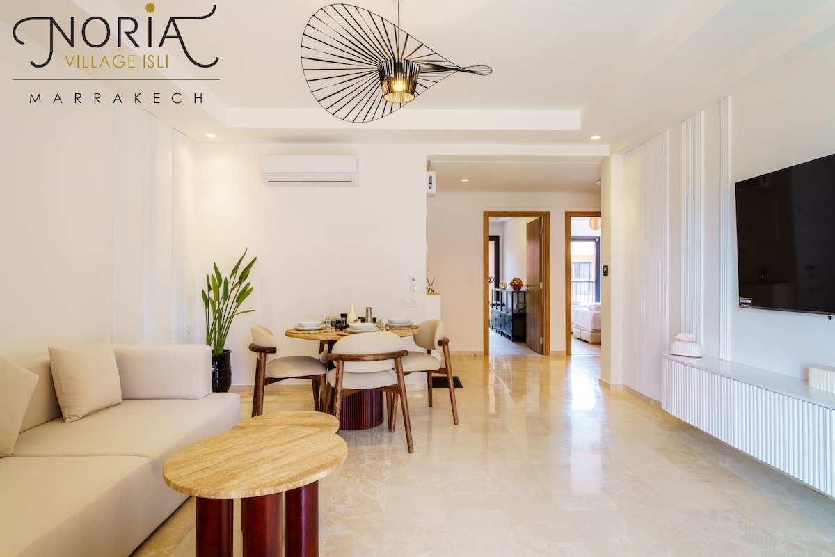 An airy living space features a round dining table surrounded by chairs, paired with a modern coffee table. A minimalist design is complemented by a wall-mounted television and a decorative light fixture above. Natural light flows through large windows, enhancing the inviting ambiance.