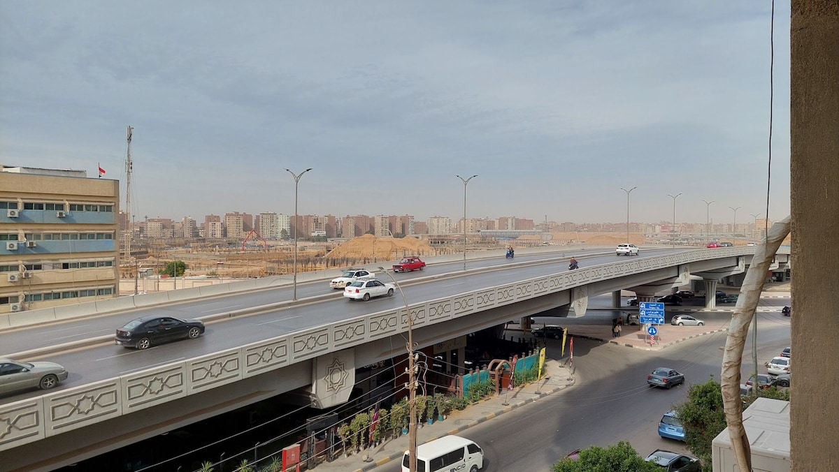 A view from an elevated position shows a busy highway with several vehicles traveling in both directions. The background features a cityscape with buildings of varying heights and distant land. A clear sky is visible, contributing to the overall sense of urban activity.