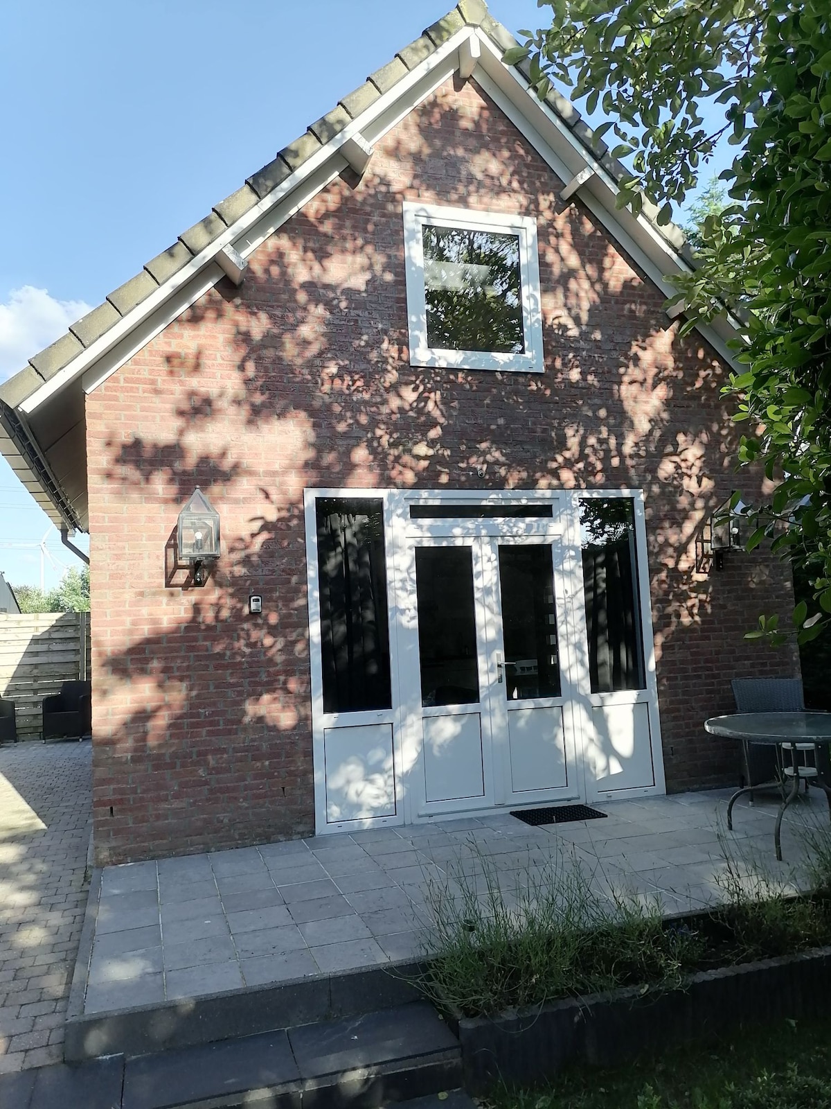 The exterior of a two-story brick house is captured, featuring a welcoming entry with large glass doors. Surrounding areas are paved with stones, and greenery is present nearby, creating a comfortable outdoor environment.