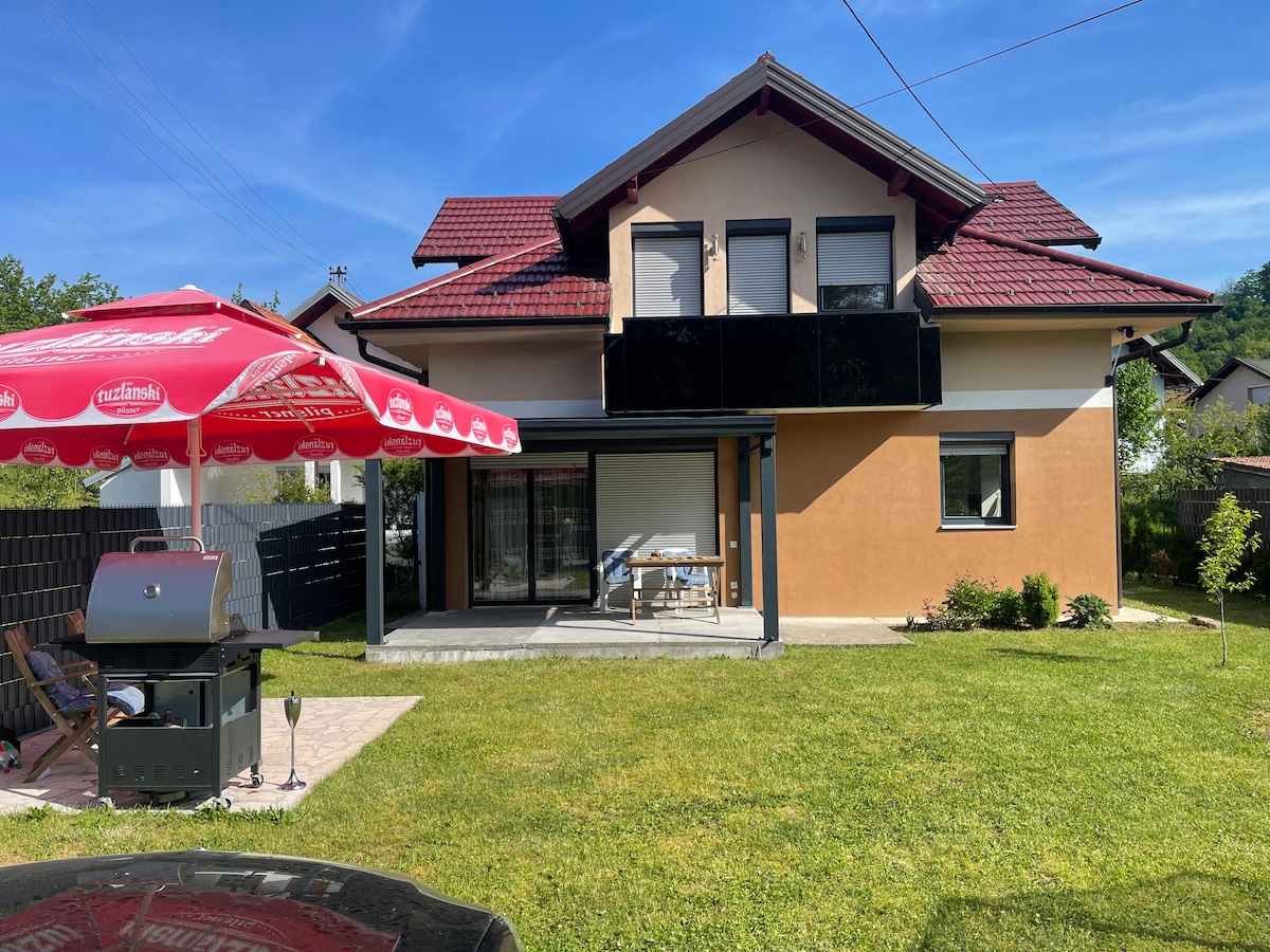 A two-story house is presented, featuring a blend of beige and black facade. An outdoor patio area is visible, with a grill and a dining table shaded by a large umbrella. A well-kept lawn surrounds the space, contributing to the inviting outdoor setting.