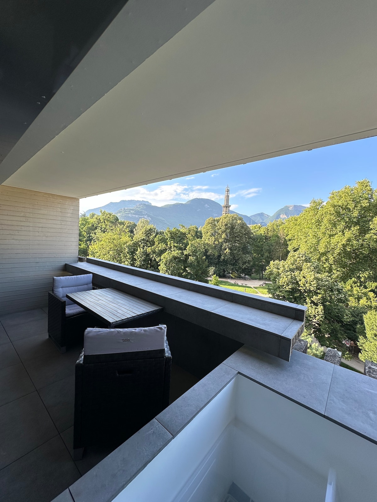 A spacious balcony features outdoor seating with a table and chairs, providing a panoramic view of green trees and distant mountains. The landscape is complemented by a clear sky, enhancing the serene atmosphere of the space.