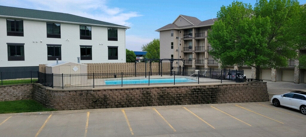 The image features a fenced outdoor pool area, complemented by a spacious deck. Surrounding greenery and nearby buildings can be seen in the background. The pool's clear water reflects the sky, creating a serene atmosphere for relaxation.