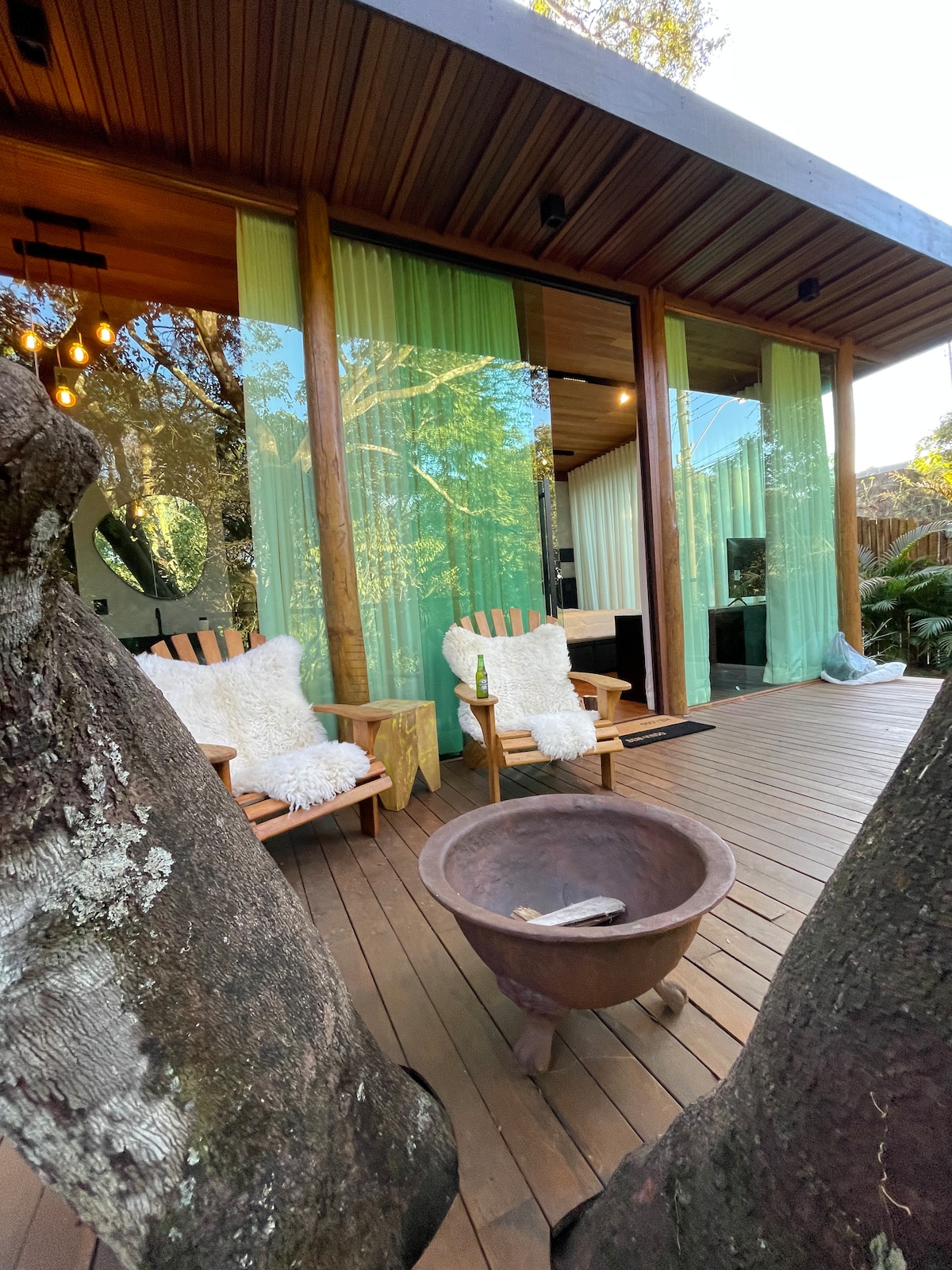 A wooden deck is shown with two relaxing chairs positioned near a fire pit. Translucent curtains hang in front of large glass doors, allowing natural light to filter into the space. Surrounding trees add to the serene atmosphere.