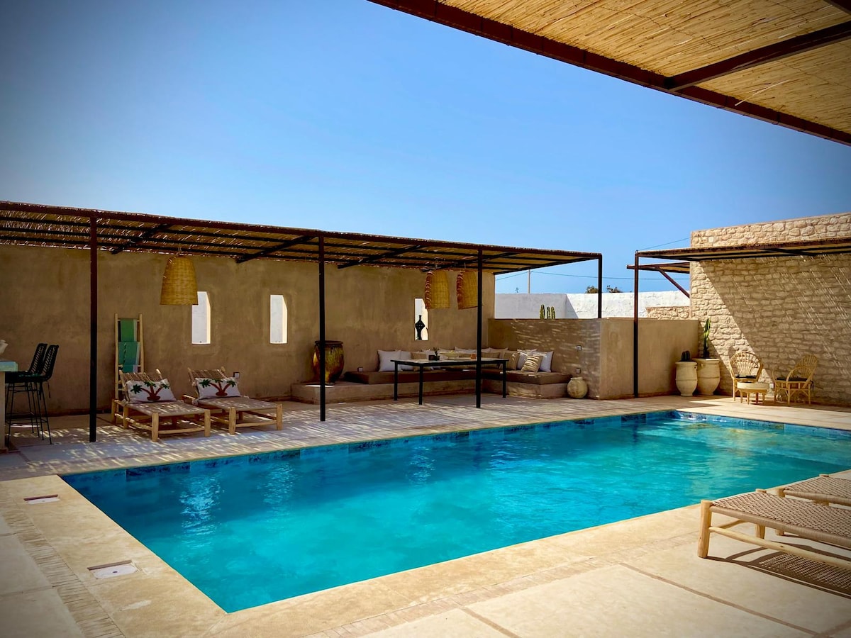 An inviting outdoor area features a swimming pool surrounded by a sun terrace. A shaded pergola offers a dining space, and comfortable seating is provided beside the pool. The villa's rustic walls and decorative elements enhance the ambience of the space.