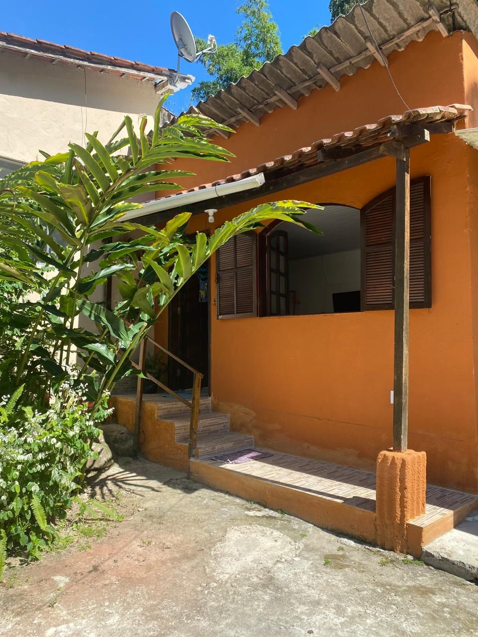 A vibrant orange exterior is displayed, featuring wooden shutters that complement the entryway. Lush green foliage frames the entrance, which is adorned with stone steps leading up to the door. A clear blue sky overhead enhances the welcoming atmosphere.